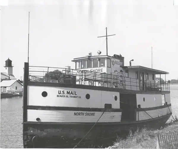 Black-and-white photograph of the mail boat North Shore moored beside a landing, with a lighthouse and buildings behind it.