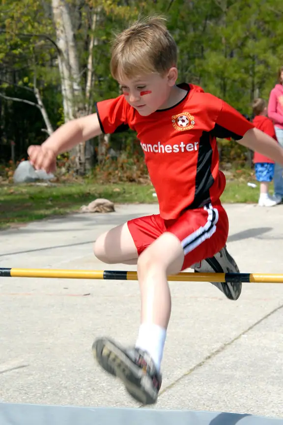 Child in a red soccer jersey jumps over a yellow-and-black hurdle outdoors.