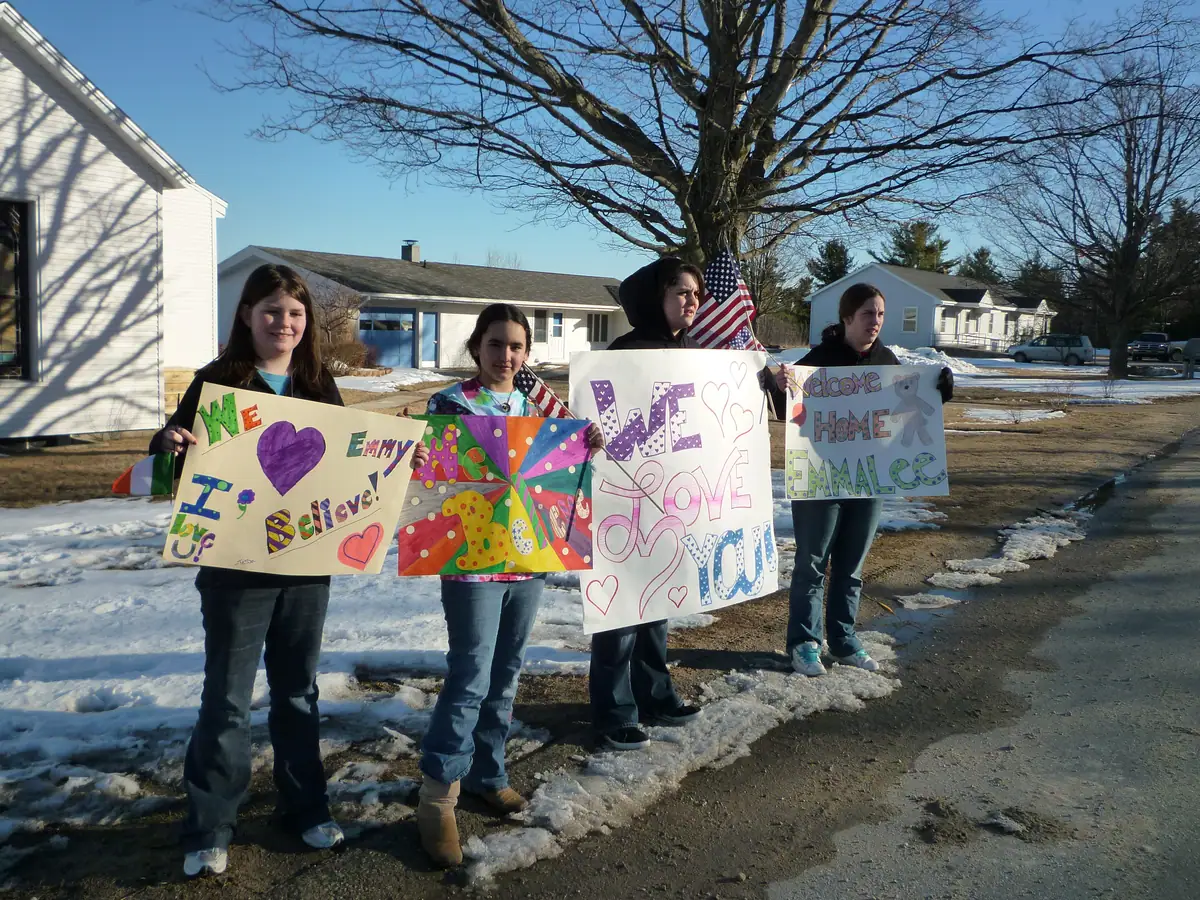 Four young people hold colorful handmade welcome signs outdoors beside homes, bare trees, and melting snow.
