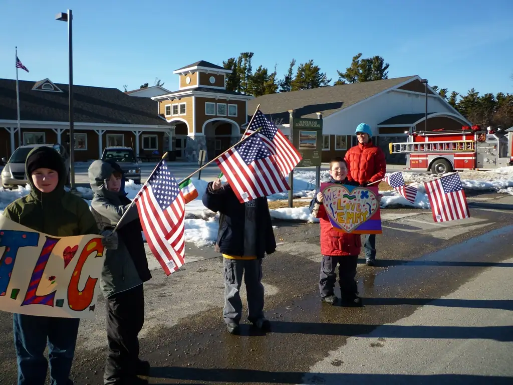 Children hold American flags and handmade signs beside a fire truck in a snowy parking area.