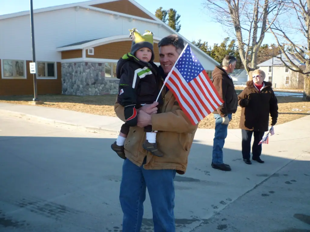 An adult carries a child and a stars-and-stripes flag outside a building while two people walk behind them.