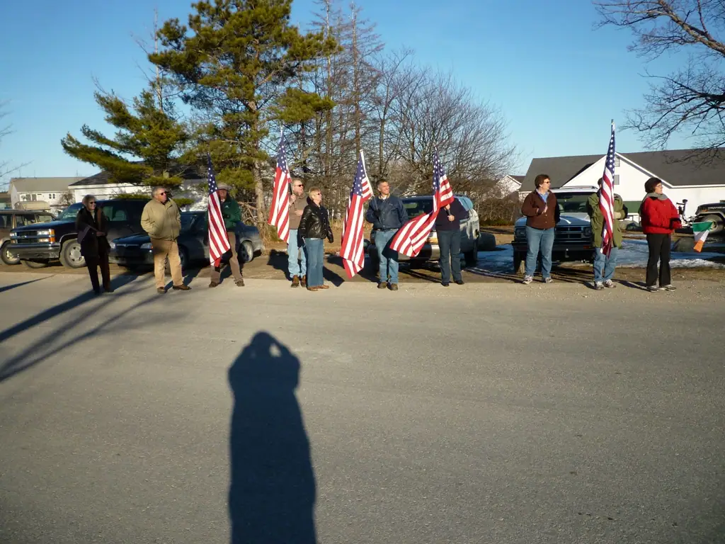 A group of people holds American flags beside parked vehicles on a sunny paved street.