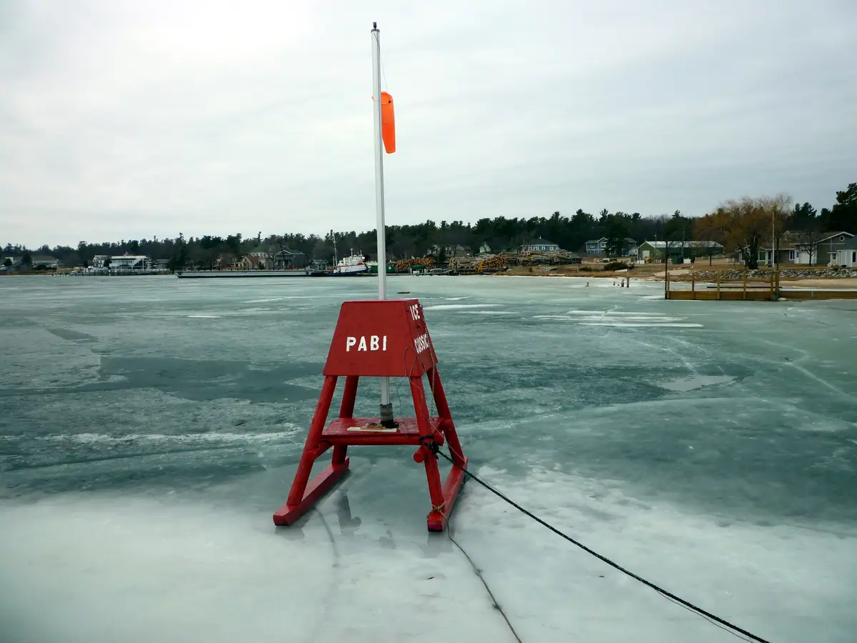 Red ice marker standing on frozen water with a shoreline, dock, boats, and houses behind it.