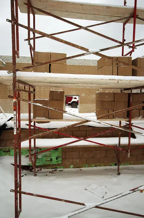 Snow-covered red scaffolding stands before a partially built concrete-block structure, with a pickup truck visible beyond it.