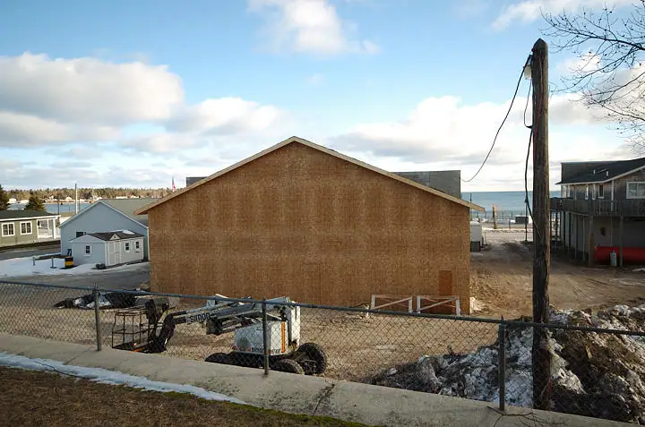 A sheathed building under construction stands behind a chain-link fence, with a skid-steer loader in front.