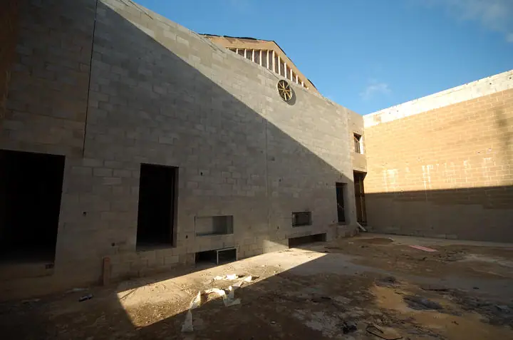 Unfinished concrete-block building interior with open doorways, window openings, roof framing, and construction debris.