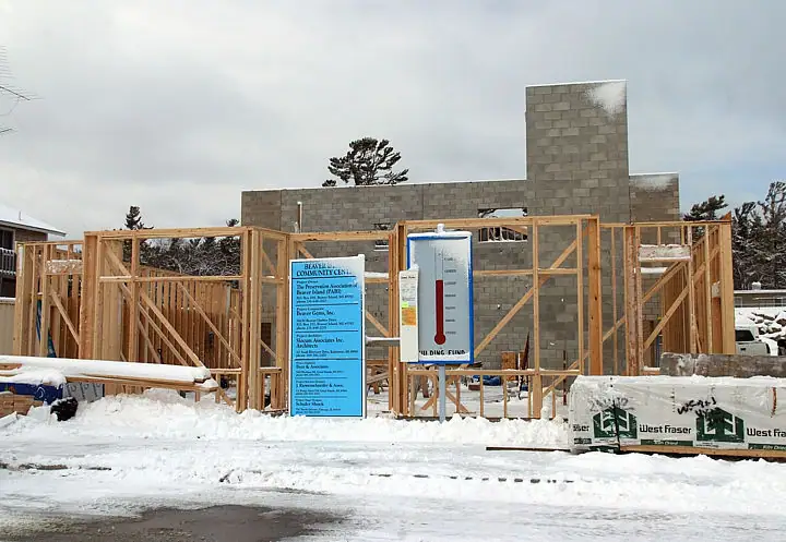 A masonry building under construction stands behind timber framing and signs in a snow-covered worksite.