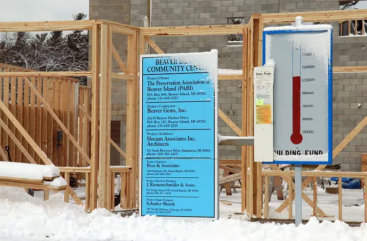 A timber-framed building rises behind project signs and a fundraising thermometer in the snow.