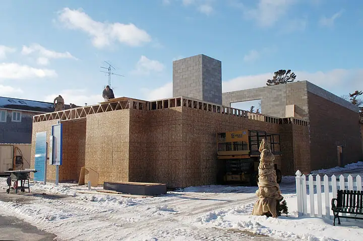 A snow-covered building under construction, with workers on the roof and a yellow loader in front.