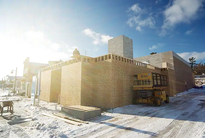 A masonry building under construction in snow, with a yellow scissor lift beside it and a worker on the roof.