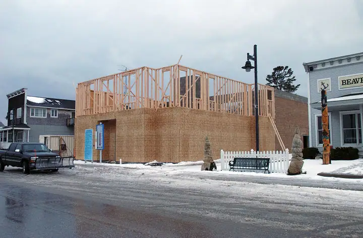 A wood-framed building under construction beside a snowy street, with a pickup truck, bench, fence, and storefront.