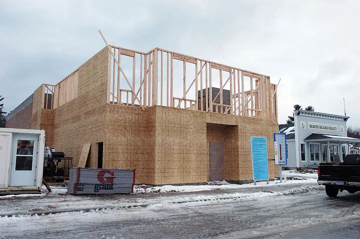A two-story wood-framed building under construction beside a snowy street, with a trailer, dumpster, and pickup truck.