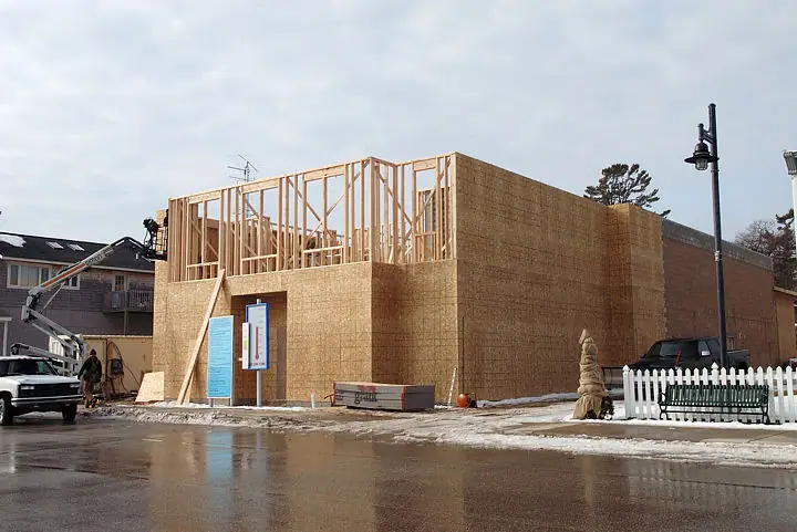 Wood-framed building under construction beside a wet street, with trucks, construction equipment, snow, and a picket fence.