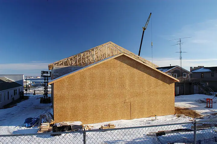 A snow-covered building under construction with exposed roof trusses, a crane, and a chain-link fence.