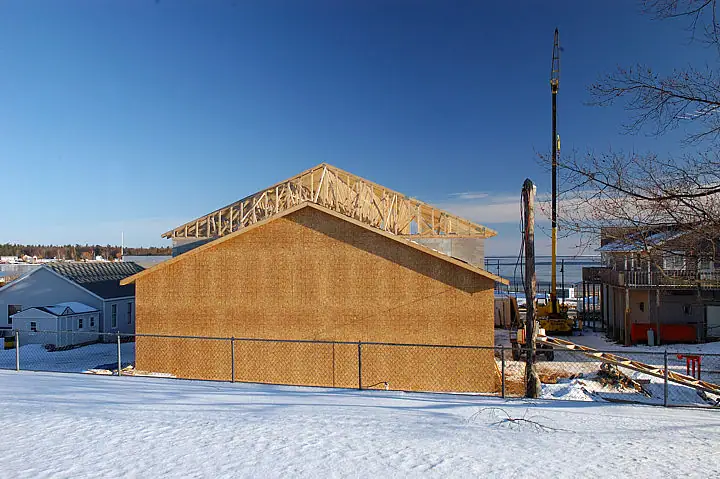 A plywood-sided building under construction stands behind a fence in a snowy setting.