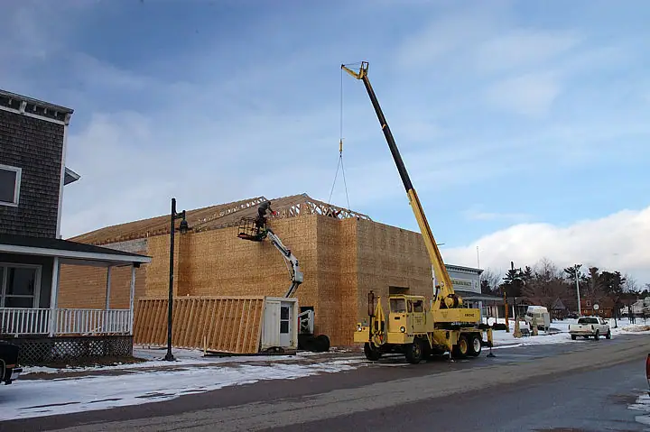 A crane lifts roof trusses beside a wood-framed building while a worker operates a boom lift.