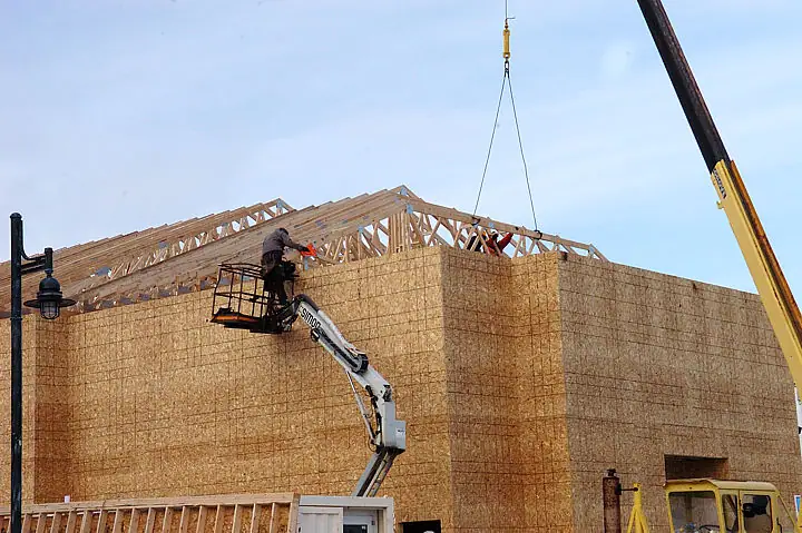 Workers use an elevated platform and crane to assemble wooden roof trusses on a sheathed building.