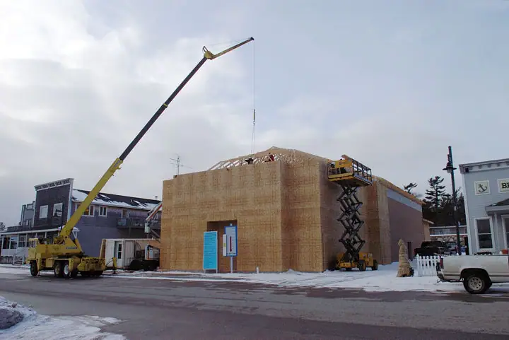 A crane lifts a roof truss beside a wood-sheathed building under construction in snowy weather.