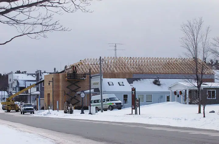 Snow-covered street beside a brick building under construction with exposed roof trusses and a yellow lift.