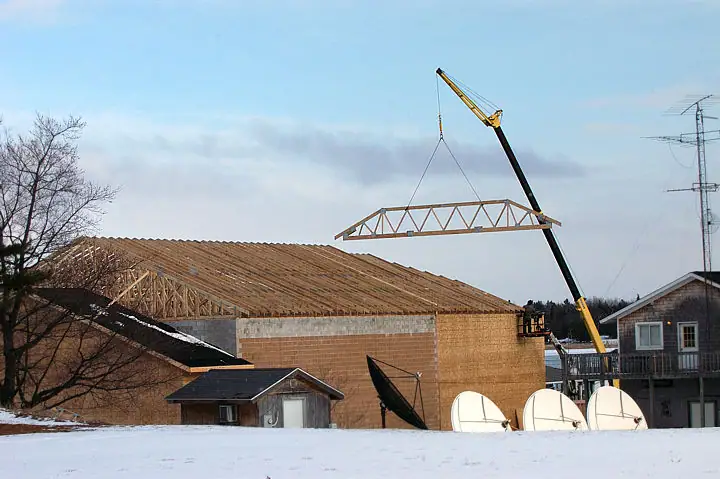 A crane lifts a wooden roof truss above a partially constructed building in snowy surroundings.