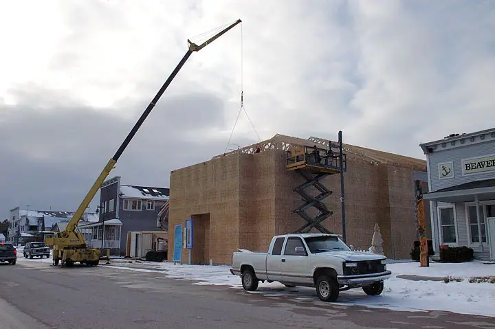 A crane lifts a roof truss over a wood-framed building beside a snowy street.