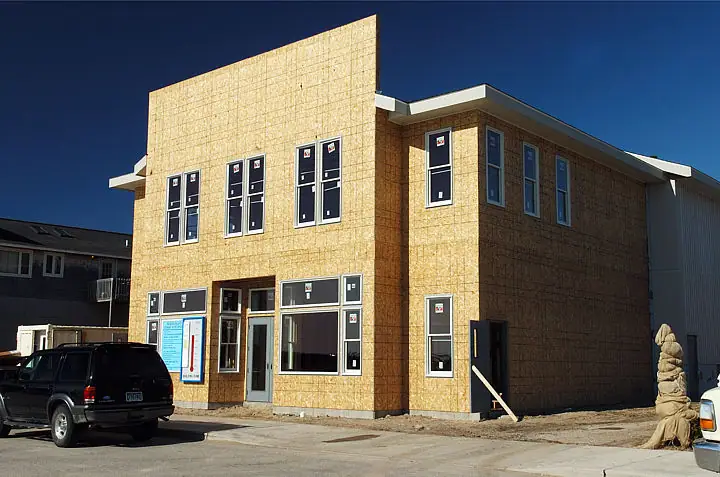 A two-story building with yellow sheathing and new windows stands under construction beside parked vehicles.