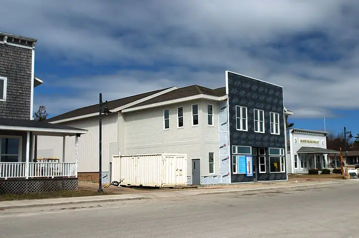 A partly finished two-story building stands beside a street, with a storage container and neighboring buildings.
