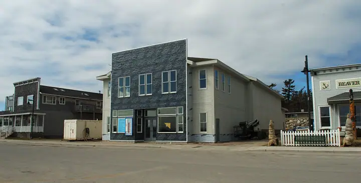 A newly built two-story building faces a broad paved street beside older buildings and a picket fence.