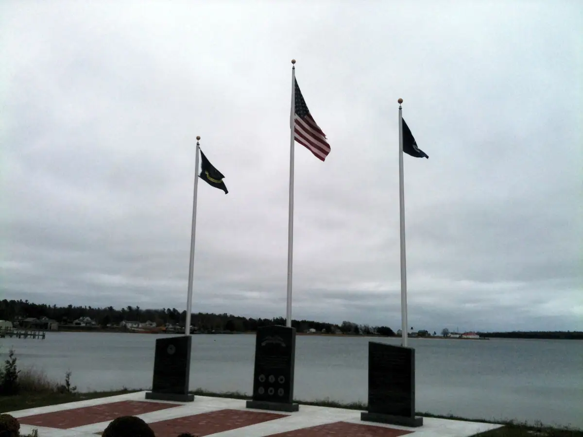 Three flagpoles and memorial plaques stand on a paved waterfront site beneath an overcast sky.