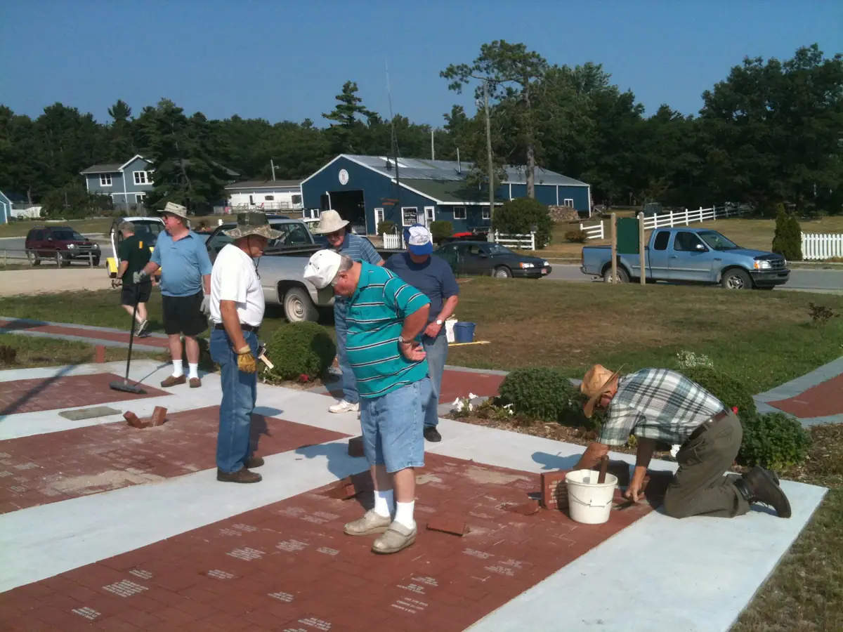 People install red brick pavers in a landscaped outdoor walkway beside a blue-sided building.