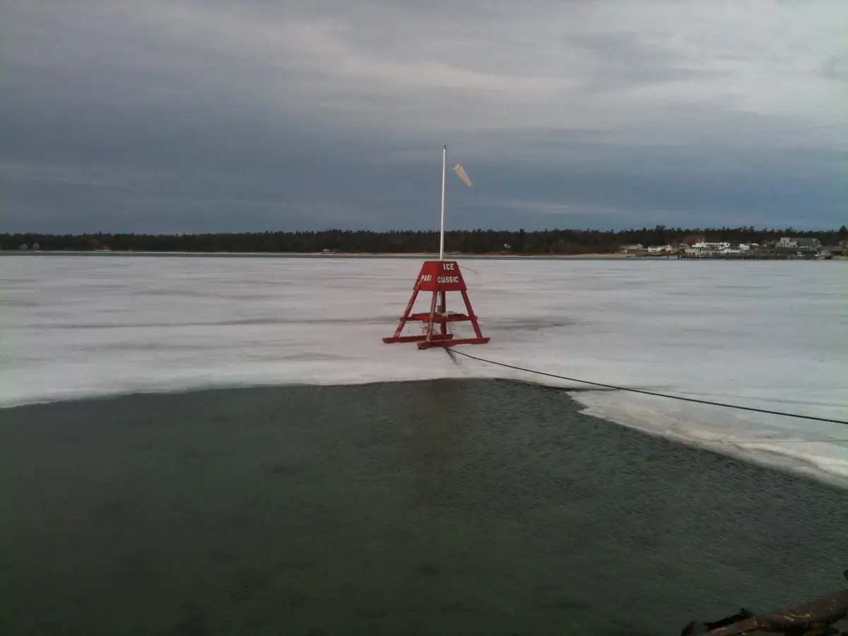 A red triangular ice marker stands on frozen water beside an open patch, with a rope extending toward the foreground.