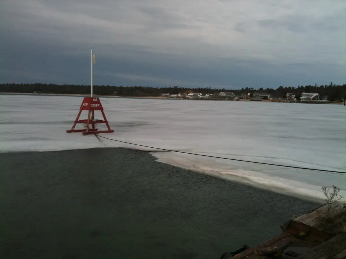A red ice marker stands on frozen water beside a dark opening, with homes and trees along the far shore.