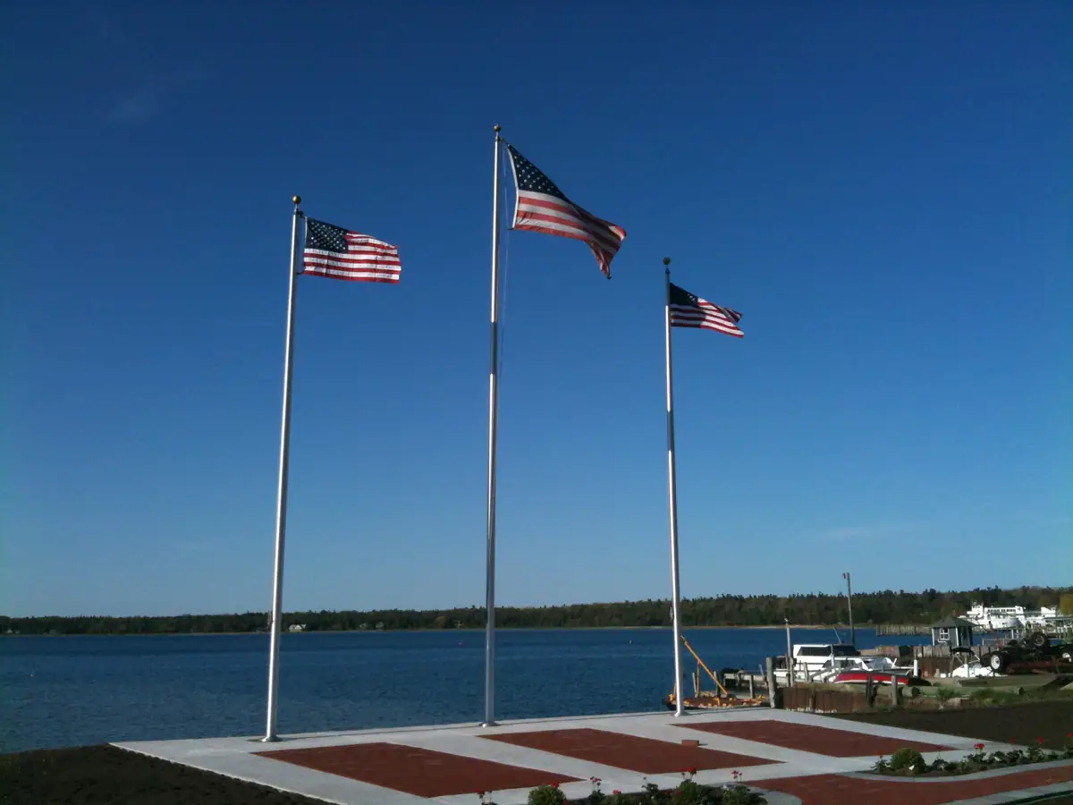 Three American flags fly above a paved waterfront plaza beside docks and boats.