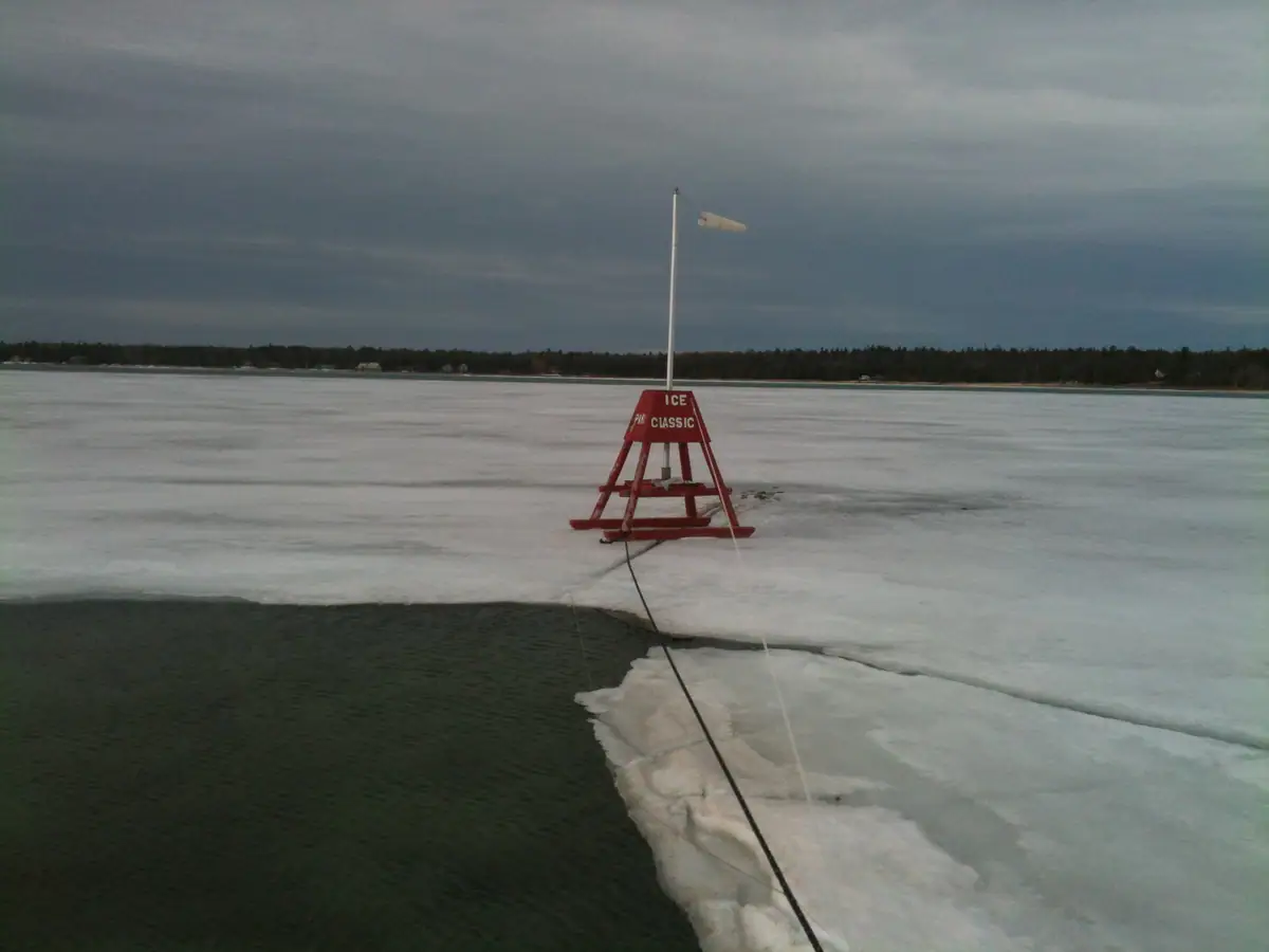 A red ice marker with a wind vane stands amid frozen water beside an opening of dark water.