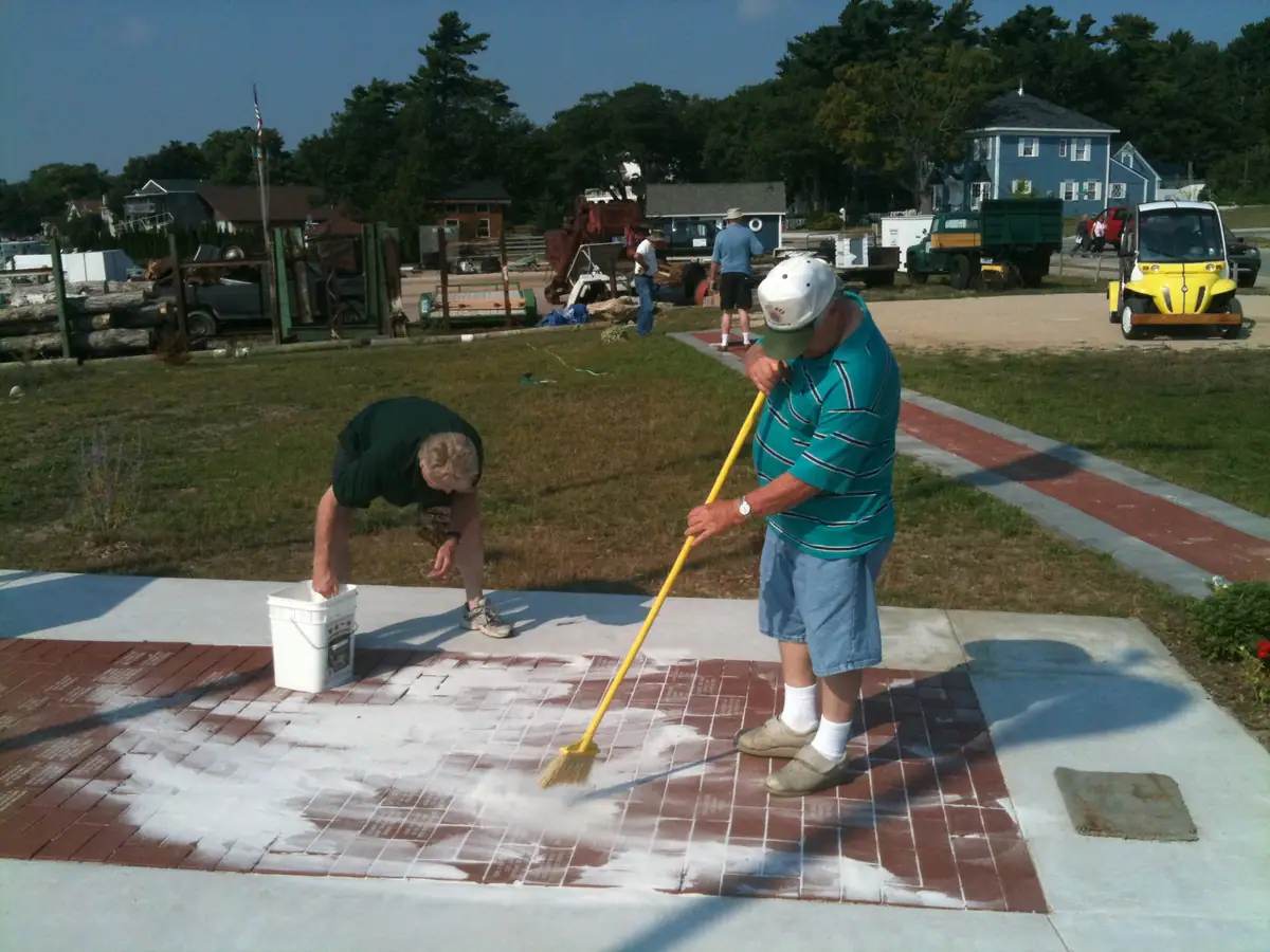 Two men clean a brick walkway with a broom and bucket while vehicles and houses stand behind them.