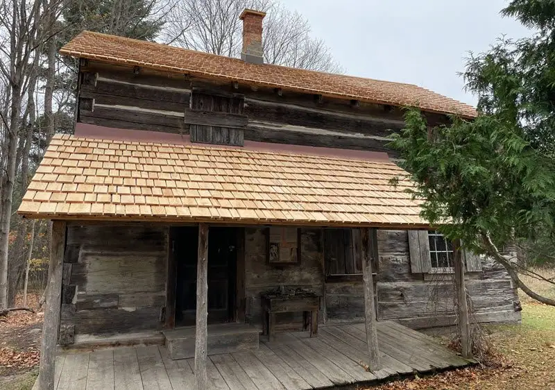 A weathered log house with a broad shingled porch stands among trees.