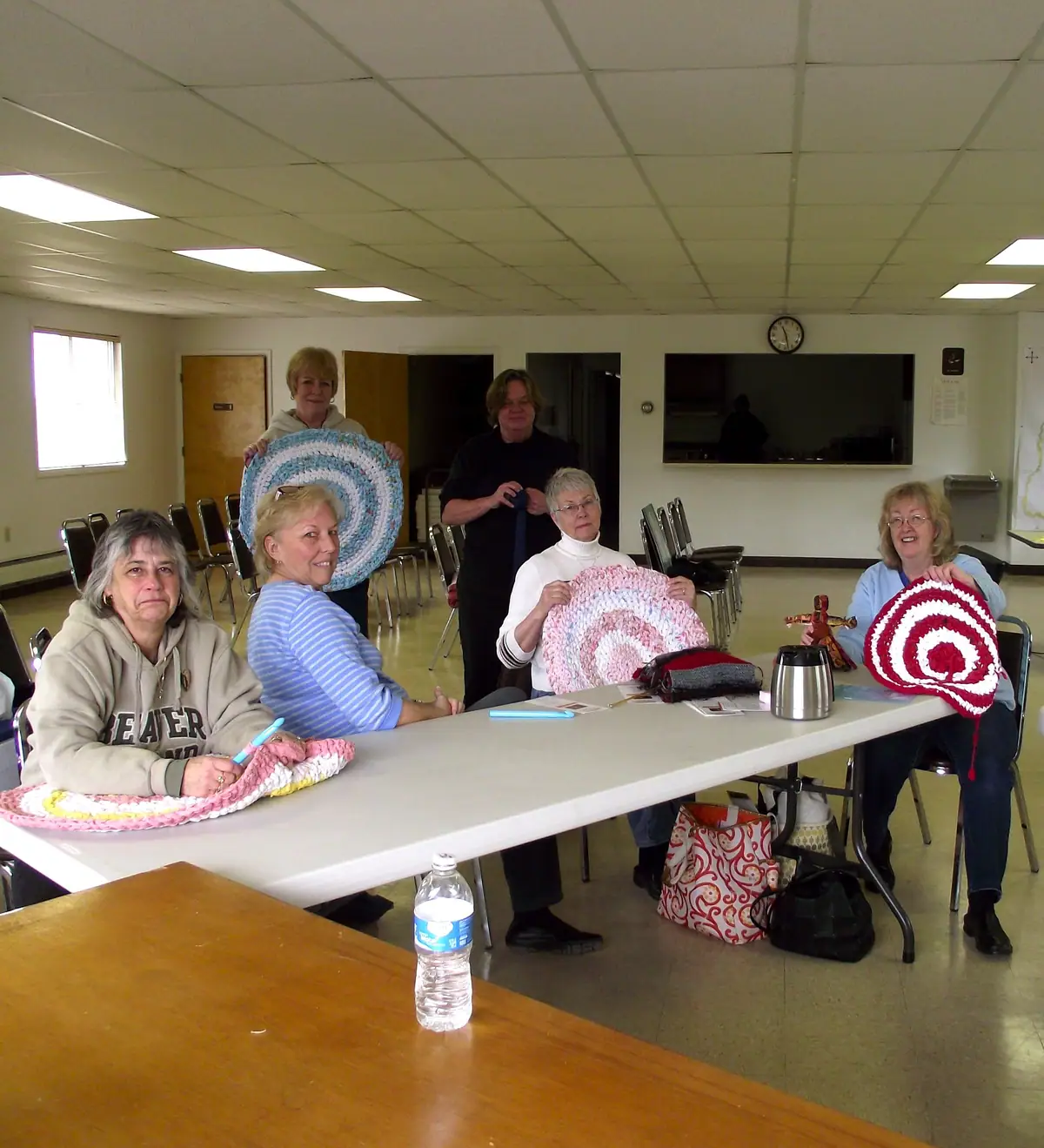 Five women display colorful circular crocheted rugs around a table in a bright meeting room.
