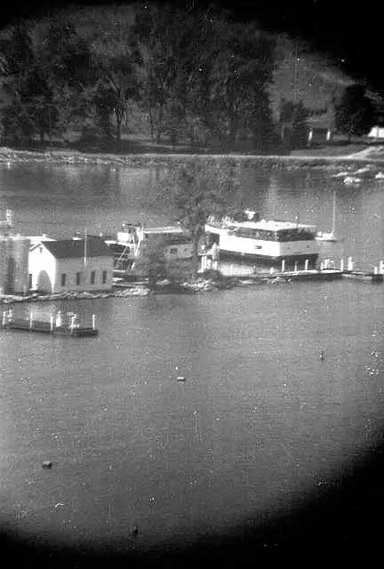 Black-and-white waterfront photograph showing a large passenger vessel, dock, small gabled building, and wooded shore.