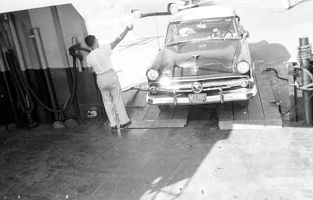A man gestures beside a vintage automobile positioned on a ferry vehicle deck.