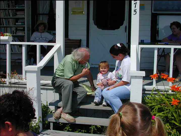 An older man, woman, and child sit on the steps of a white porch surrounded by flowers and onlookers.