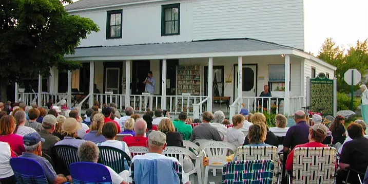 A crowd seated in lawn chairs listens to a speaker on the porch of a white building.