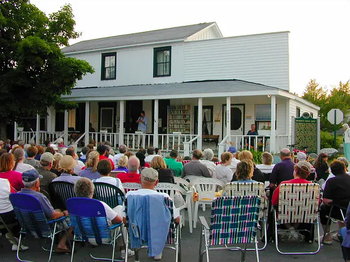 A large outdoor audience faces a speaker on the porch of a white building.