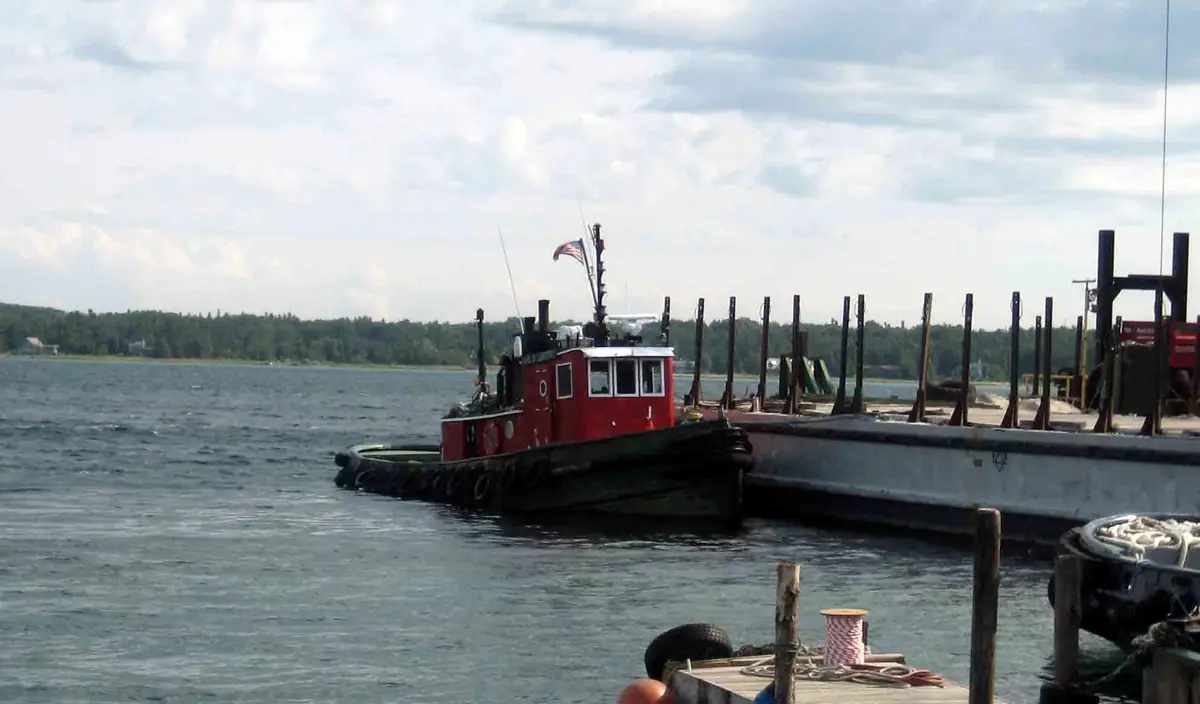 Red tugboat beside a barge and dock, with a wooded shoreline across the water.