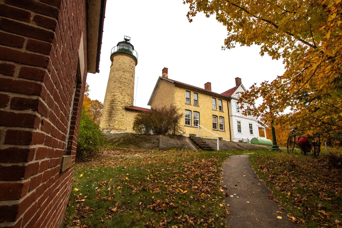 A brick lighthouse and adjoining yellow-brick dwelling stand beside a paved path beneath autumn foliage.