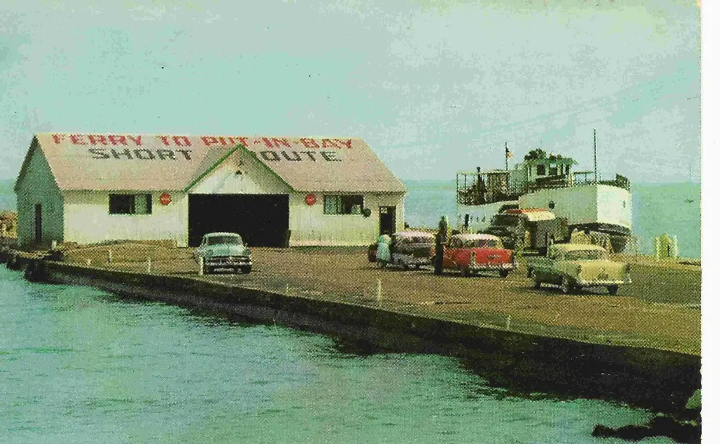 A ferry beside a pier and terminal building with vintage cars and a large roof sign.