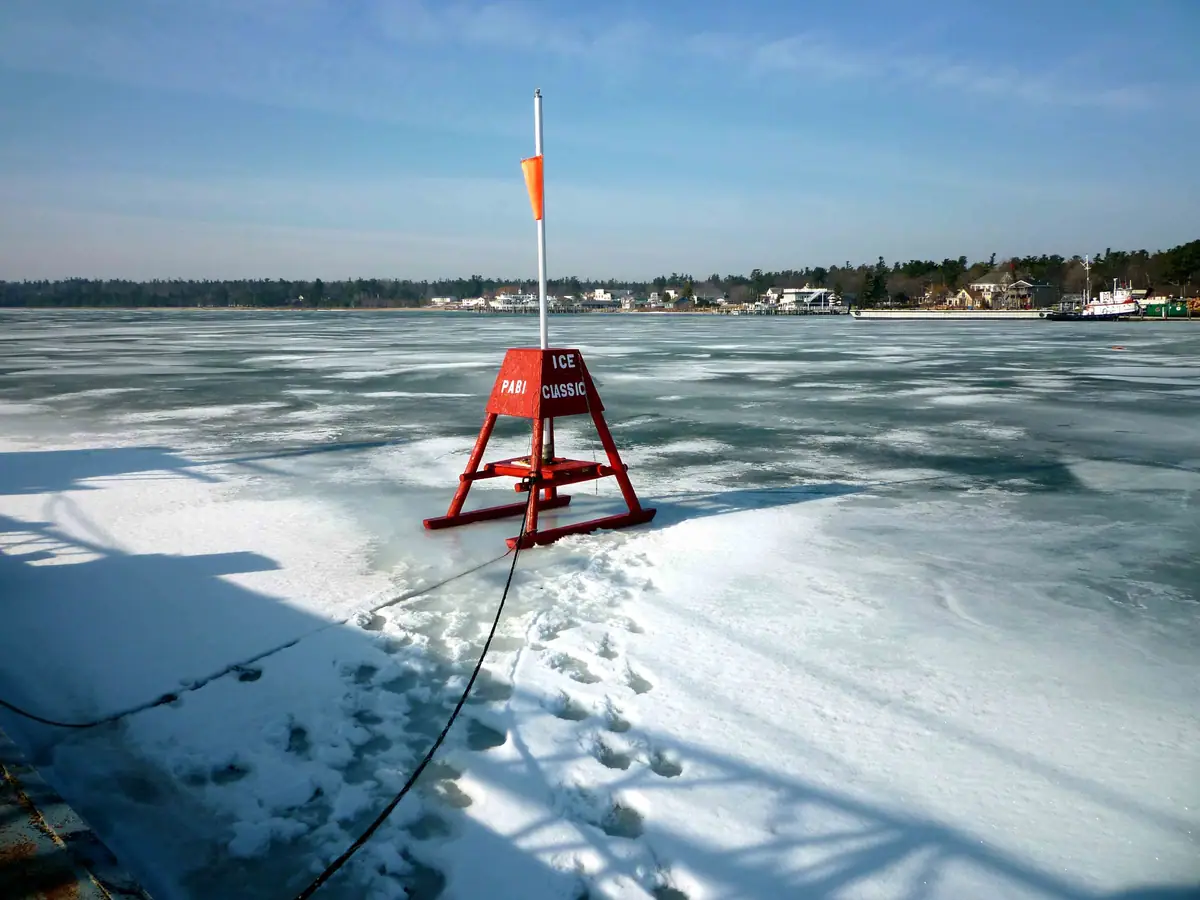 A red ice marker stands on frozen water, with a shoreline settlement and boats in the distance.
