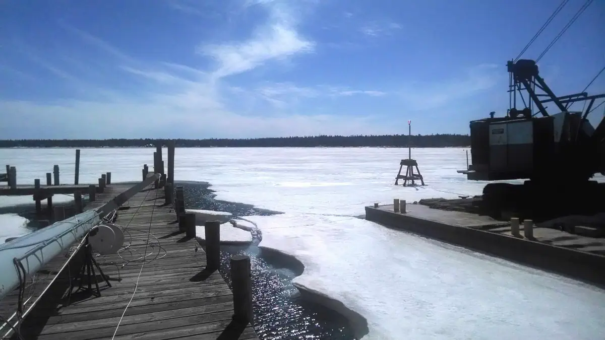 A wooden pier and industrial equipment beside ice-covered water under a bright blue sky.