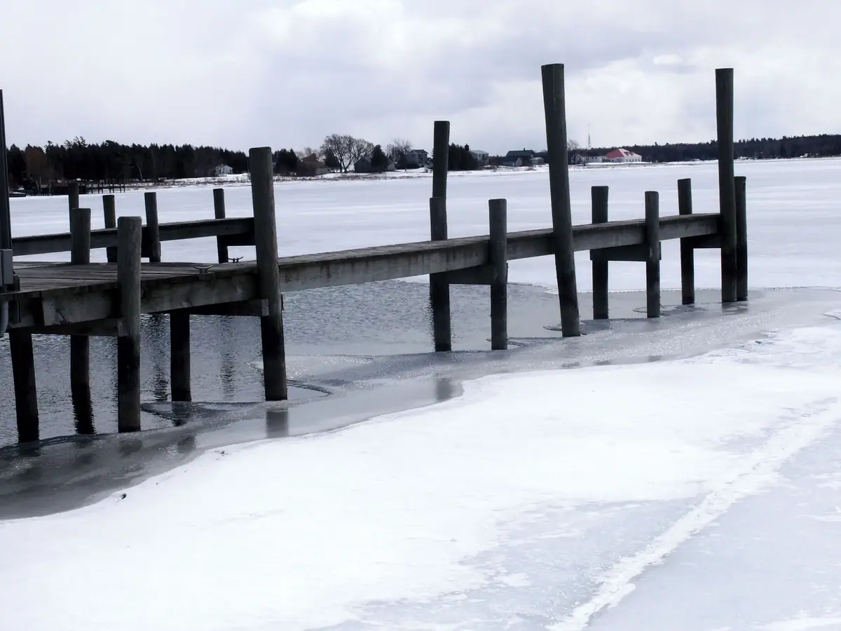 A weathered wooden pier crosses partially thawed ice toward a tree-lined shore with houses.