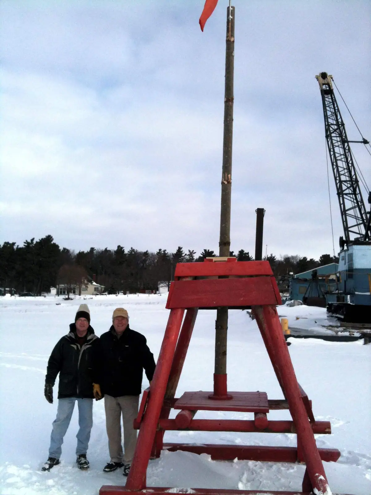 Two people stand beside a red wooden frame and flagpole in a snowy work area with a crane behind them.