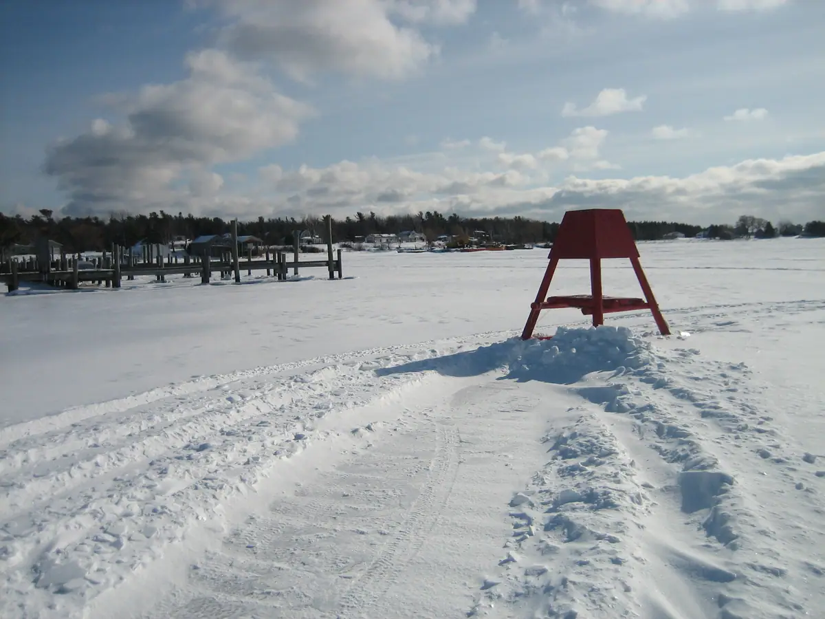 A red A-frame tower stands on a snowy frozen surface beside a dock, with houses and trees in the distance.
